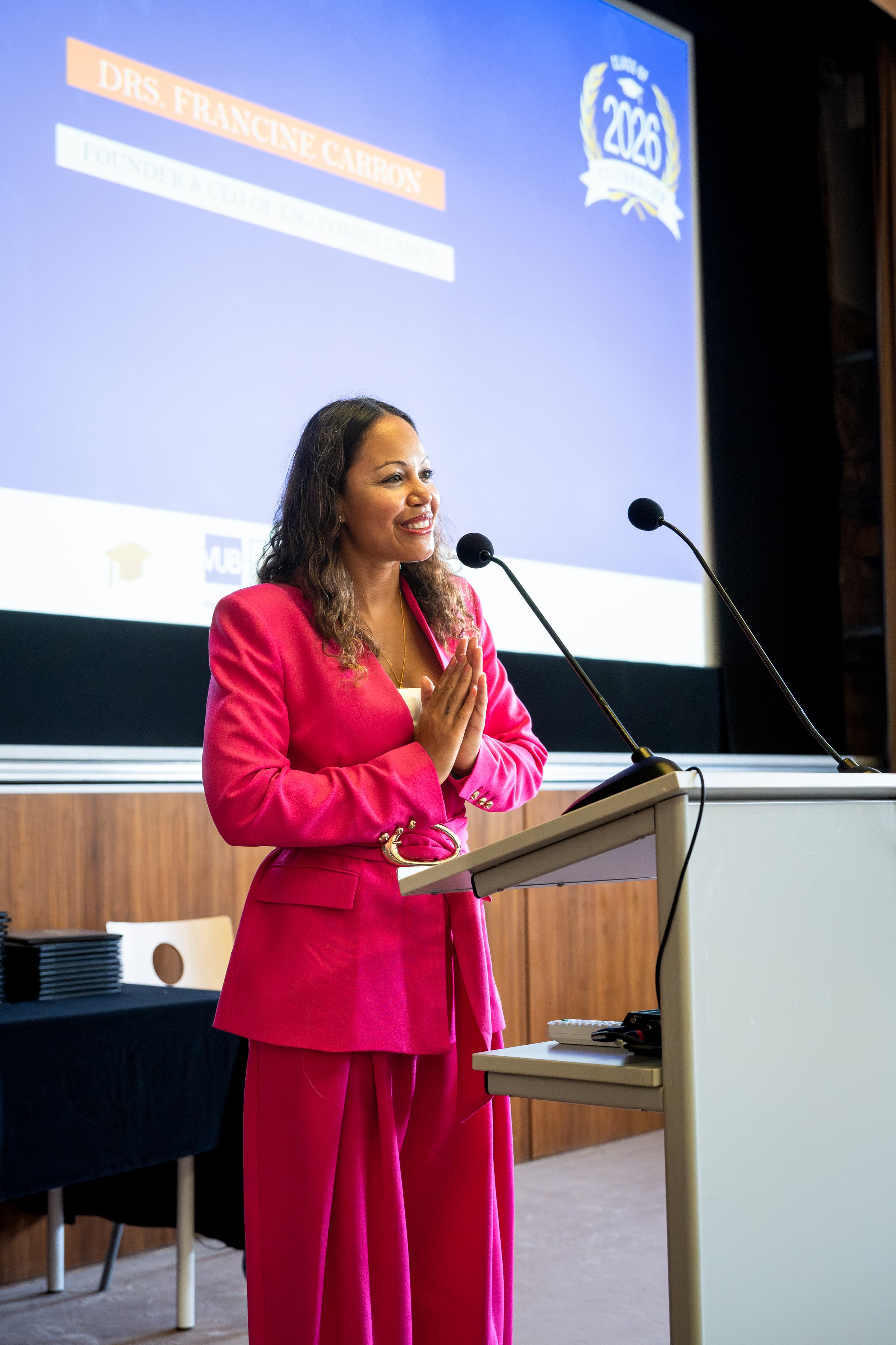 Woman in a vibrant pink suit speaks at a podium with her hands clasped together.
