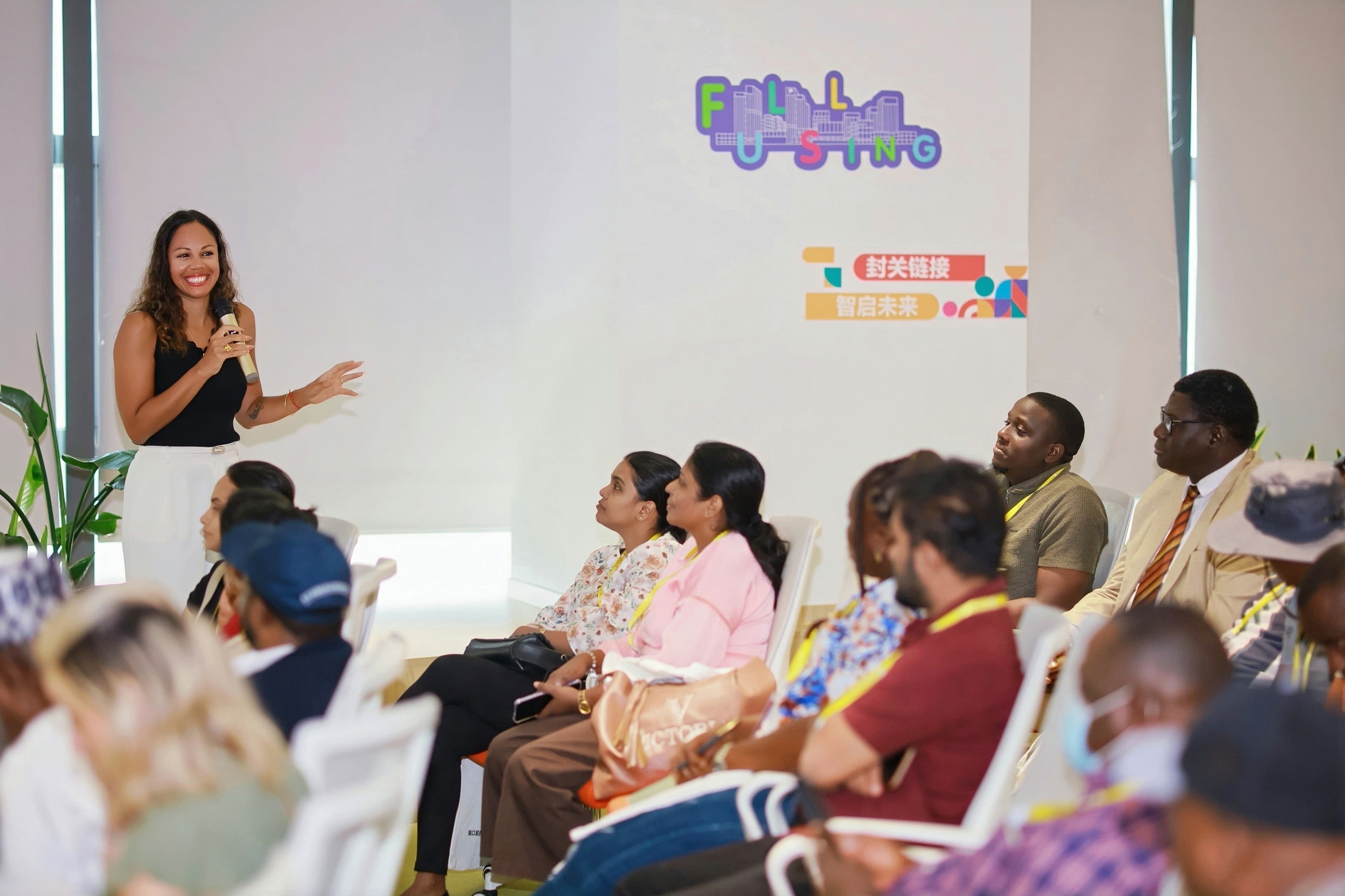 Smiling woman presents with a microphone to a diverse seated audience in a bright room.
