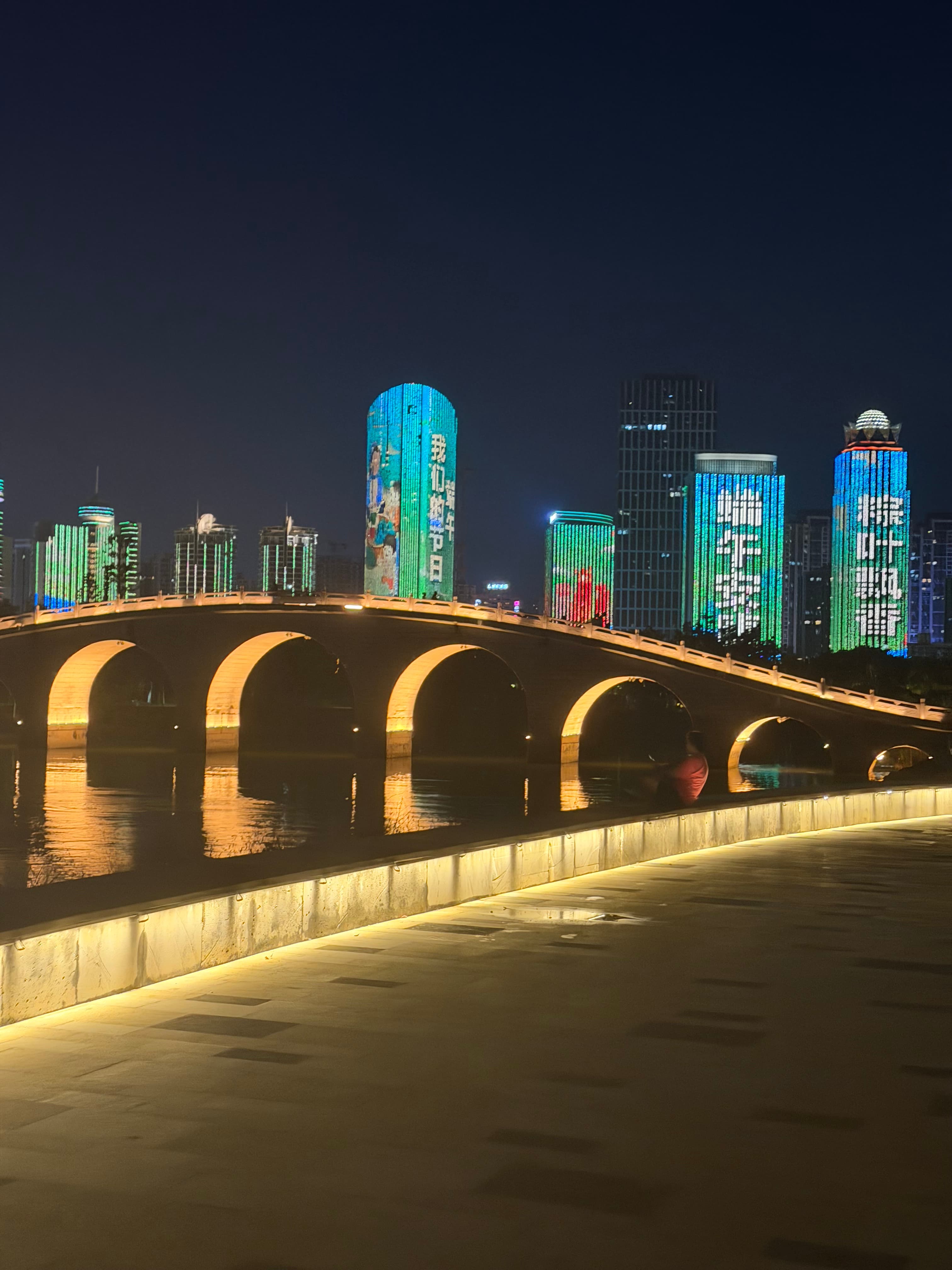 Illuminated stone arch bridge reflecting in water with vibrant LED-lit skyscrapers in the background.