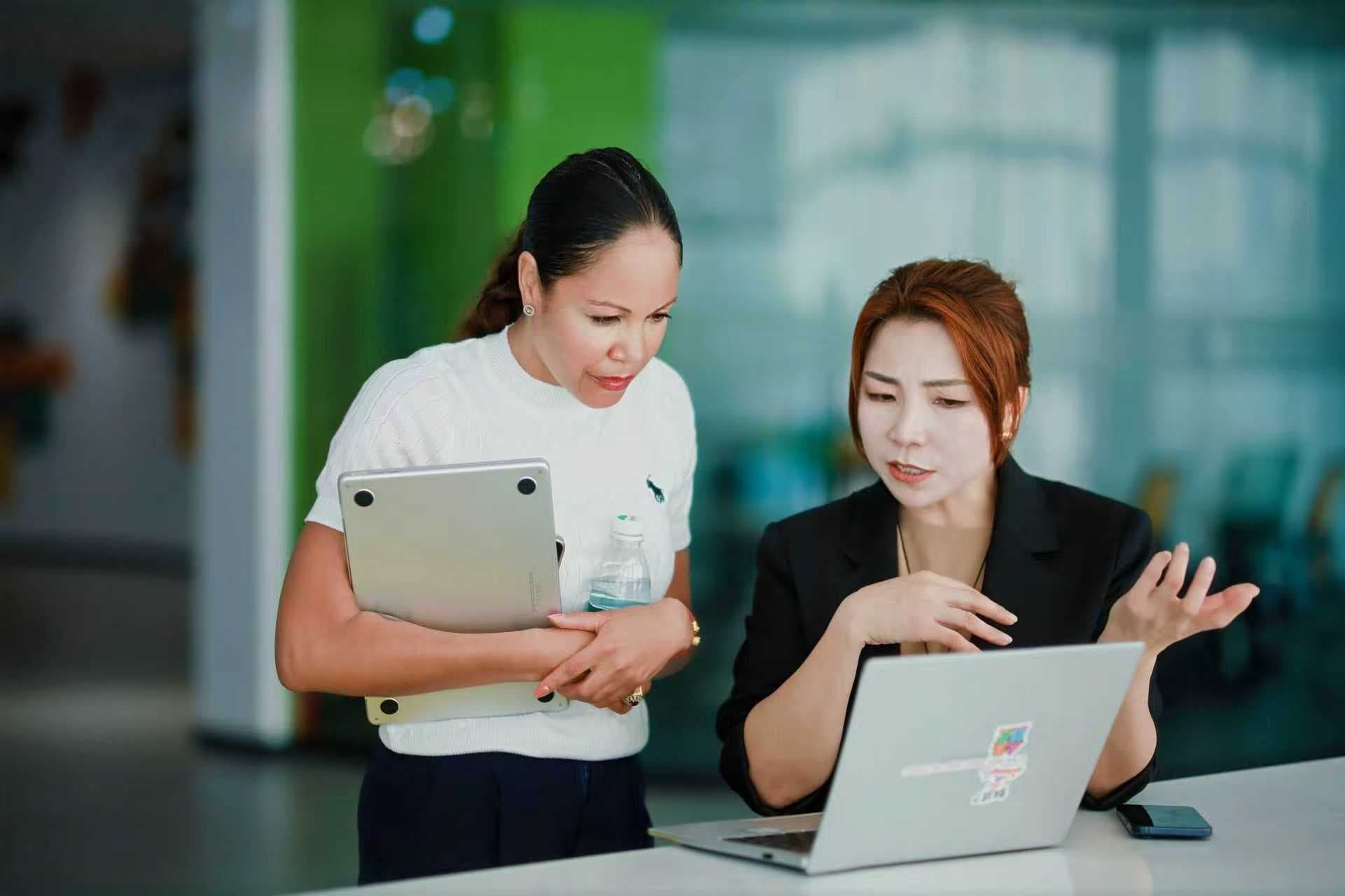 Two professional women collaborate while looking intently at a laptop screen in a modern office.