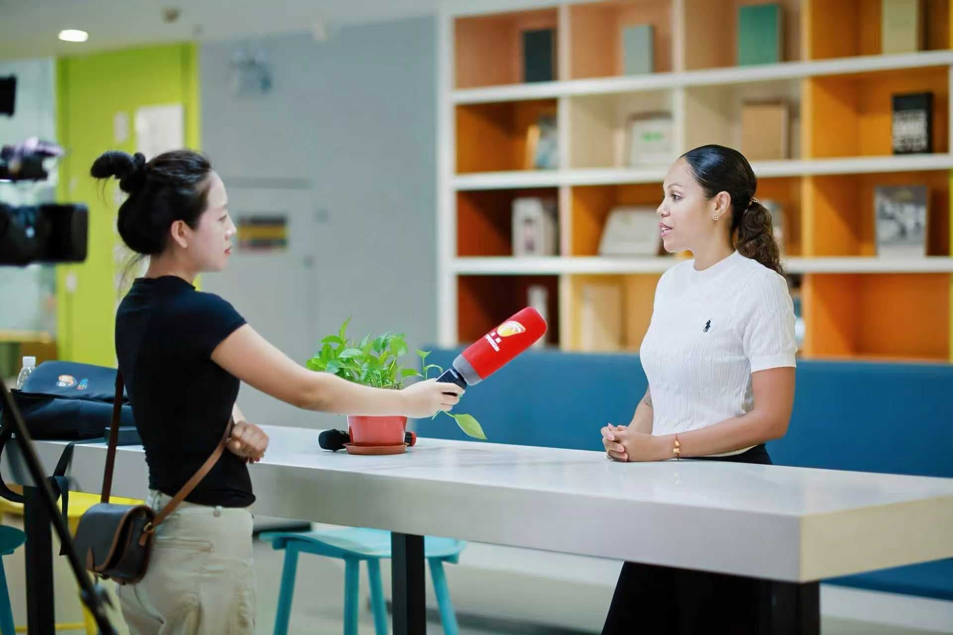 Woman in black interviews another woman with a red microphone in a modern office.